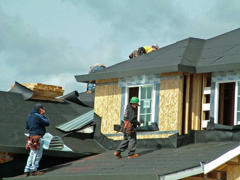 Homeowner examining roof shingles and gutters from the ground beside a residential house.